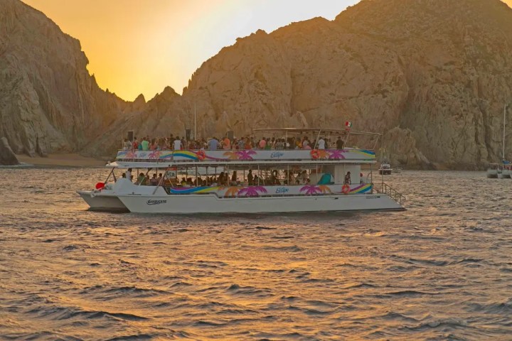 Crowded boat on ocean at sunset near rocky cliffs.