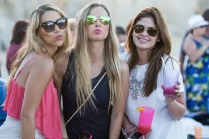 Three women in sunglasses pose outdoors with drinks at a casual gathering.