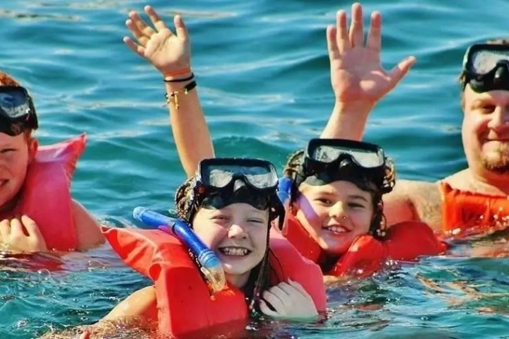 Four people snorkeling in the ocean wearing red life vests and goggles, waving and smiling.