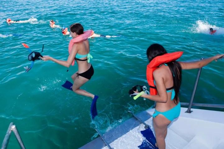 People snorkeling, jumping from boat into turquoise water, wearing life vests and fins.