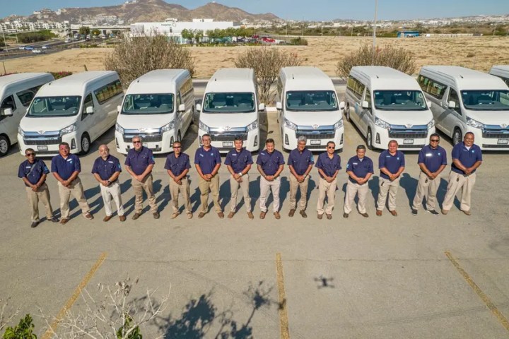 Twelve people in blue shirts stand in front of a row of white vans in a parking lot.